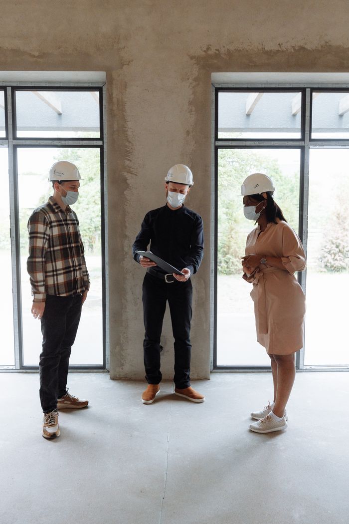 about-01 Three people in masks and hard hats discuss real estate indoors with natural lighting.